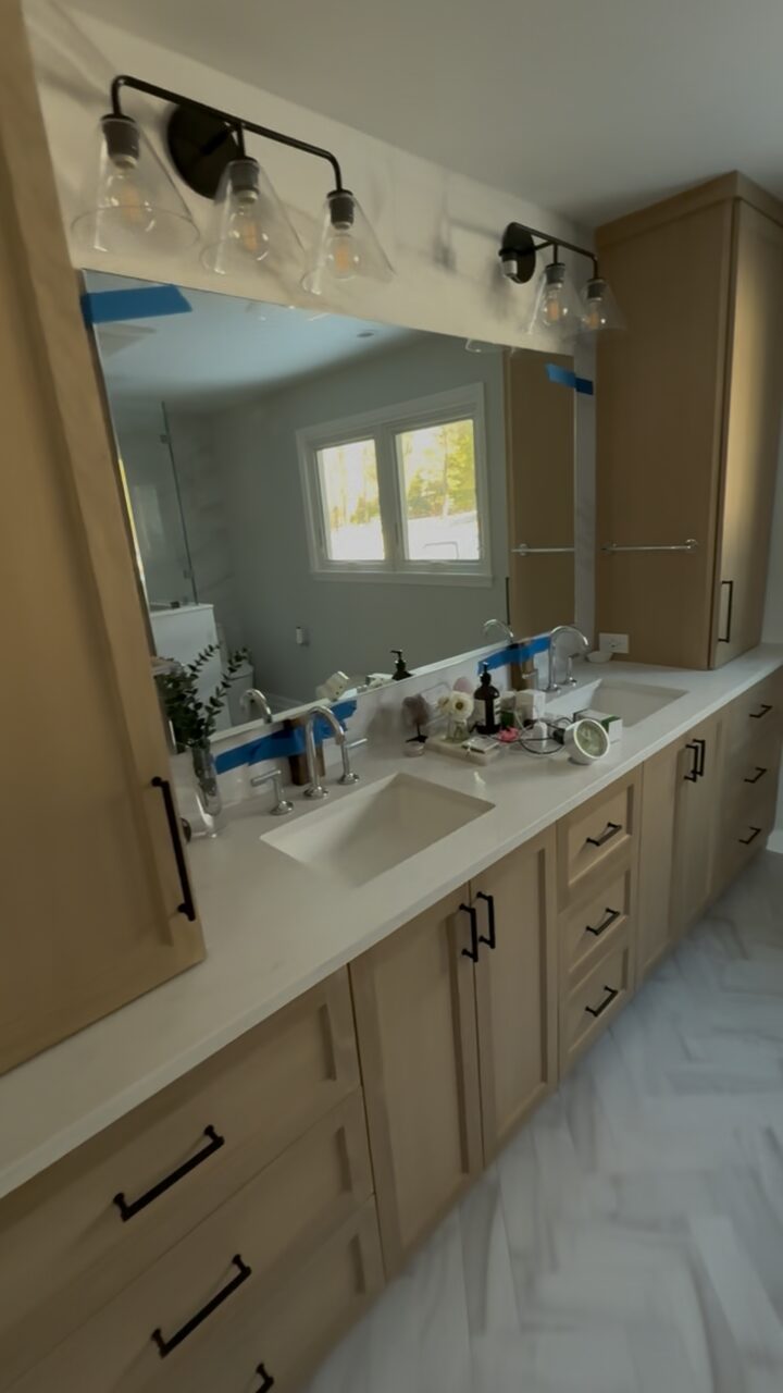 White oak double vanity with a quartz countertop, black hardware and a herringbone marble floor.